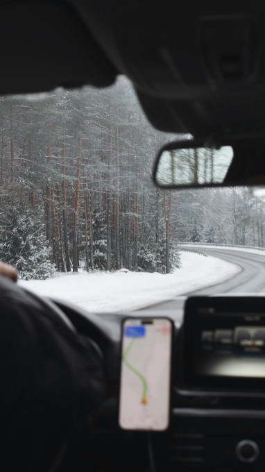 View from a car interior showing a snowy road surrounded by tall trees, with a GPS navigation screen on the dashboard.