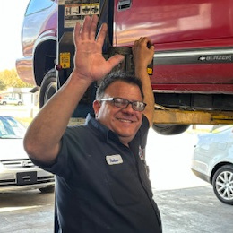 Roine Del Risco, Automotive Technician of AAMCO in South Tampa, beside a lift in the shops garage, waving hello